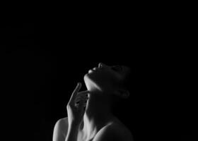 A black and white portrait of a woman with her head tilted upwards, touching her neck with her hand. The background is dark, creating a dramatic contrast, copy space above photo