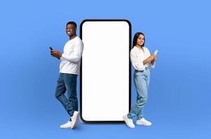 On a deep blue background, a multiracial couple, both smiling, stands adjacent to an enormous smartphone with a blank display, ready for showcasing apps or designs. photo