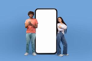A young Hindu man and woman are standing next to a blank screen against a blue background. They are posing confidently, looking directly at the camera. photo
