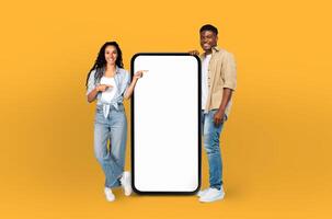 A young man and woman stand in front of a bright yellow background, both pointing at a blank screen. They appear engaged and focused on the screen, possibly presenting or discussing photo