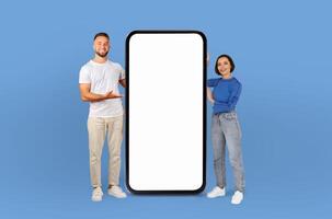A man and woman are standing side by side, holding up a blank screen against a solid blue background. Their hands are steady as they display the empty screen to the viewer. photo