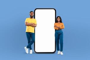 Black couple positioned next to a blank screen while standing against a blue background. They appear to be engaged in a presentation or discussion, with the screen ready for content to be displayed. photo