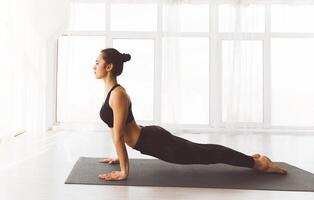 In a sunlit studio with large windows, lady is engaged in a yoga practice, focusing on an upward-facing dog pose, a common back-bending exercise in various traditions of yoga photo