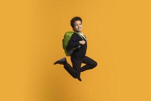 African American boy wearing a black blazer and black pants is jumping in front of a bright yellow background. He has a green backpack on and is smiling at the camera. photo