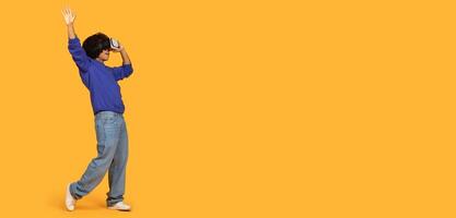 Guy is energetically dancing while wearing a blue long-sleeve shirt and light blue jeans. He is raising one hand and smiling as he moves against a vibrant yellow backdrop, using VR headset, copy space photo