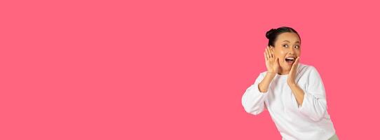 A joyful Asian lady stands against a vibrant pink backdrop, enthusiastically gesturing as if sharing exciting news, panorama with copy space photo