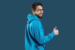 un hombre vistiendo un azul capucha es mostrado en el imagen, levantamiento su mano a dar un pulgares arriba gesto. el mans cara es no visible, y él aparece a ser en pie en contra un llanura antecedentes. foto