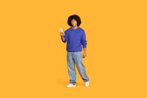 A young man stands confidently with a smartphone in hand, wearing a blue sweater and light jeans. He has a cheerful expression and is positioned in front of a bright yellow background photo
