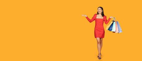 A woman in a red dress is standing in front of a yellow background. She is holding a stack of colorful shopping bags in her right hand and gesturing with her left hand, panorama with copy space photo