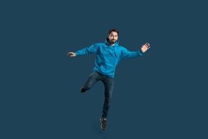 A young man wearing a vibrant blue hoodie is playfully balancing on one leg, smiling widely. The setting features a simple dark background, emphasizing his dynamic pose photo
