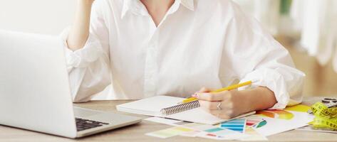 A woman sits at a desk, writing in a notebook with a pencil. She has a laptop in front of her and color swatches on the table. photo