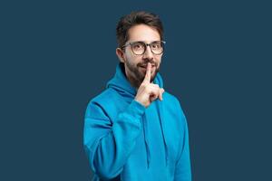 A man in a bright blue hoodie stands against a matching blue background, placing one finger over his lips as if to gesture for silence or secrecy. photo
