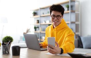 Business And Telecommuting Concept. Portrait of smiling young asian man using smartphone and modern laptop computer, reading text message, sitting at desk with gadgets, working at home office photo