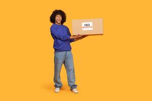 A cheerful guy with curly hair stands against a vibrant orange backdrop, joyfully holding a large brown box marked with FREE. The man showcases a wide smile, embodying excitement and anticipation photo