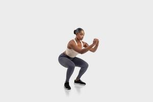 Black woman with dark hair pulled back in a bun wears a white tank top and grey leggings as she performs a squat exercise. Her fists are clenched at her chest and she is looking straight ahead photo