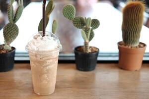 Fresco hielo latté frapé café con bulbo en el plastico vaso en de madera mesa y cactus planta Decorar en mostrador en cafetería. Copiar espacio foto