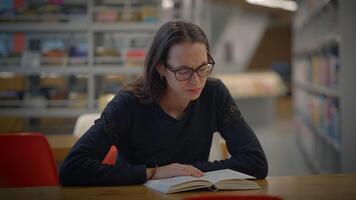 A focused student is deeply engaged in reading a book in the library while wearing glasses video