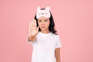 Dissatisfied Asian woman in white pajamas and sleeping mask at home with stop gesture with palms on home pastel pink background. Lifestyle concept relax good mood. photo