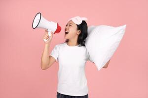 Sad and crazy Asian woman in pajamas and blindfold hugging a pillow. She is screaming into a sound amplifier on pastel pink background in studio. Upset. photo