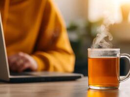 Steaming cup of tea beside a laptop, cozy workspace in warm light. photo