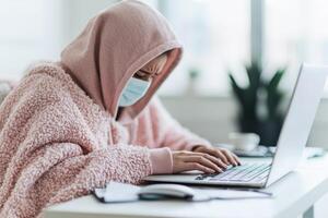 Person in a cozy hoodie, focused on a laptop, working or studying at home with a mask on. photo
