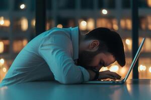 Man stressed at work with head down on laptop, dimly lit office background. photo