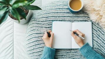 Person writing in notebook beside coffee and plant on cozy blanket. photo