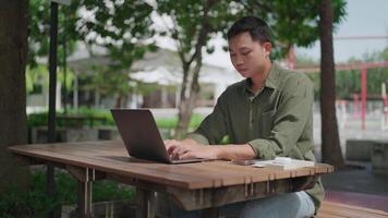 Asian man multitasking outdoors in a park using laptop and smartphone while working on documents on wooden table. Phone has incoming call while working. Freelance, remotely, sitting in city park. video