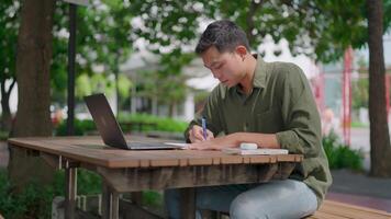 Study and take notes. Asian man is studying online, writing notes while looking at laptop screen on outdoor table in park. Working remotely during daytime. Daytime. Summer. In nature. Study outdoors. video