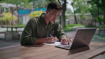 Using laptop to work and take notes. Asian man is working by taking notes in document and working by looking at data on laptop placed on table. Outdoor park. Freelance work. Deadline. Researching. video