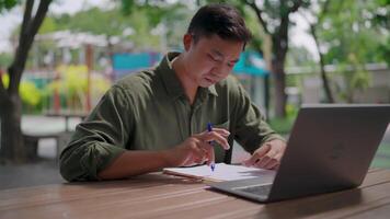Asian man writing a document and editing something in the document. Working hard and stressed. Outdoor in a park during the day. Laptop and documents on the table. Deadline. Working outdoors concept. video