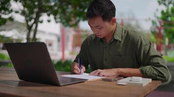 Asian man working on document, laptop and phone next to each other on the same table. Man taking notes, outdoors in a park, researching and writing document, freelance work, texting on phone, gadget. video