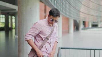 Student standing answering a phone call with bag on his shoulder. Asian man standing talking on the phone with bag on his shoulder leaning against the railing of a university building. Communication. video