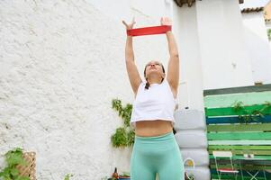 Young woman exercising with resistance band outdoors in sunny courtyard, practicing fitness routine and stretching photo