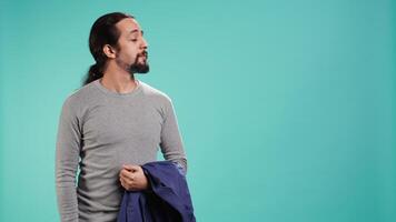 Man holding jacket in one hand while looking around, contemplating, creating a sense of curiosity. Person holding coat showing curiosity for surroundings, studio background, camera B video