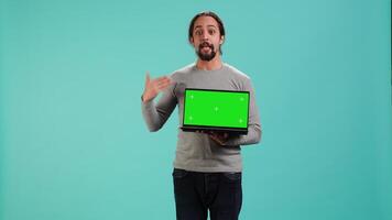 Tech content creator doing influencer marketing using green screen laptop, isolated over studio background. Arab man holding empty copy space mockup notebook, doing review, camera A video