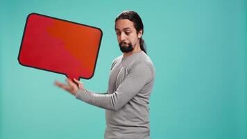 Smiling man holding red speech bubble sign of empty copy space for message. Joyous person presenting thought bubble cardboard used as promotion concept, studio background, camera B video