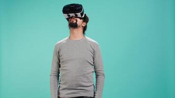 Excited tech fan wearing virtual reality goggles, looking around. Arab man using high tech futuristic modern VR glasses, making tactile touching gesturing, studio background, camera B video