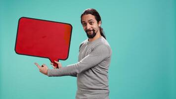 Smiling man holding red speech bubble sign of empty copy space for message. Joyous person presenting thought bubble cardboard used as promotion concept, studio background, camera B video