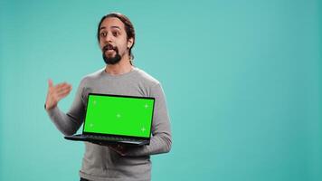 Tech content creator doing influencer marketing using green screen laptop, isolated over studio background. Arab man holding empty copy space mockup notebook, doing review, camera B video