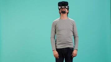 Excited tech fan wearing virtual reality goggles, looking around. Arab man using high tech futuristic modern VR glasses, making tactile touching gesturing, studio background, camera A video