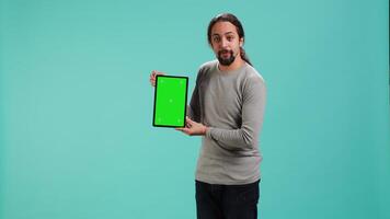 Jolly man presenting on green screen tablet, isolated over studio background. Upbeat person holding blank copy space chroma key device used for showcasing ads, camera A video