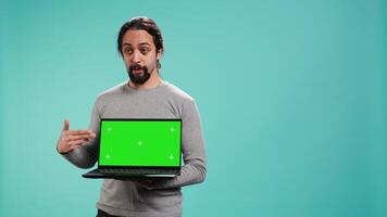 Man presenting green screen mobile laptop, isolated over studio background. Arab person holding copy space chroma key notebook device used for advertising brands, camera B video