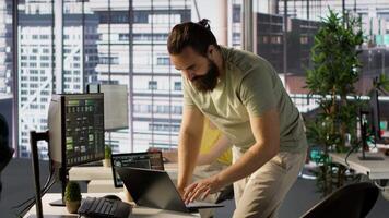 Admin at office desk utilizing cloud computing resources on laptop to train large scale machine learning models. Man monitoring company systems using notebook while standing in workspace, camera A video