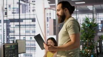 Cheerful admin holding laptop chatting with colleagues while working with machine learning tech. Man transforming data into predictive models using notebook and talking with coworkers, camera A video