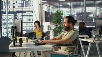 IT worker with disability sitting in wheelchair in office working at computer desk, focused on testing programs. Software engineer with paraplegia in inclusive workplace checking code on PC, camera B video