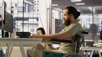 IT worker with disability sitting in wheelchair in office working at computer desk, focused on testing programs. Software engineer with paraplegia in inclusive workplace checking code on PC, camera A video