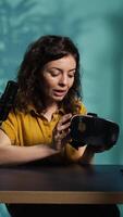 Vertical Reviewer discussing with viewers about high tech VR headset using smartphone on holder to capture footage. Woman chats with fans during stream, filming virtual reality goggles with phone, camera B video
