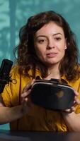Vertical Reviewer discussing with viewers about high tech VR headset using smartphone on holder to capture footage. Woman chats with fans during stream, filming virtual reality goggles with phone, camera A video