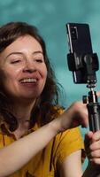 Vertical Smiling woman in studio using smartphone on selfie stick to record , greeting subscribers. Joyous entertainer hosts q and a session with viewers using cellphone, saluting fans, camera A video